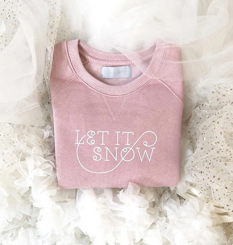 a pink Bob & Blossom sweatshirt with a test graphic reading let it snow surrounded by white tutus